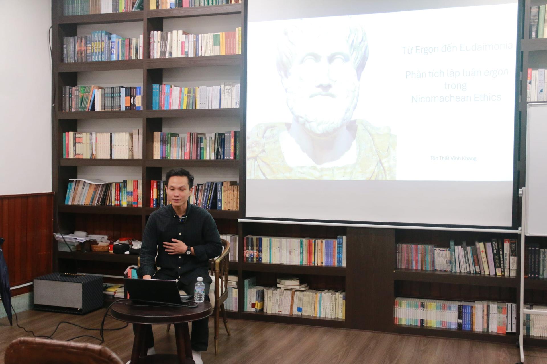 TS Tôn Thất Vĩnh Khang ngồi trình bày dưới màn chiếu có tượng bán thân Aristotle và tựa đề «Từ Ergon đến Eudaimonia».