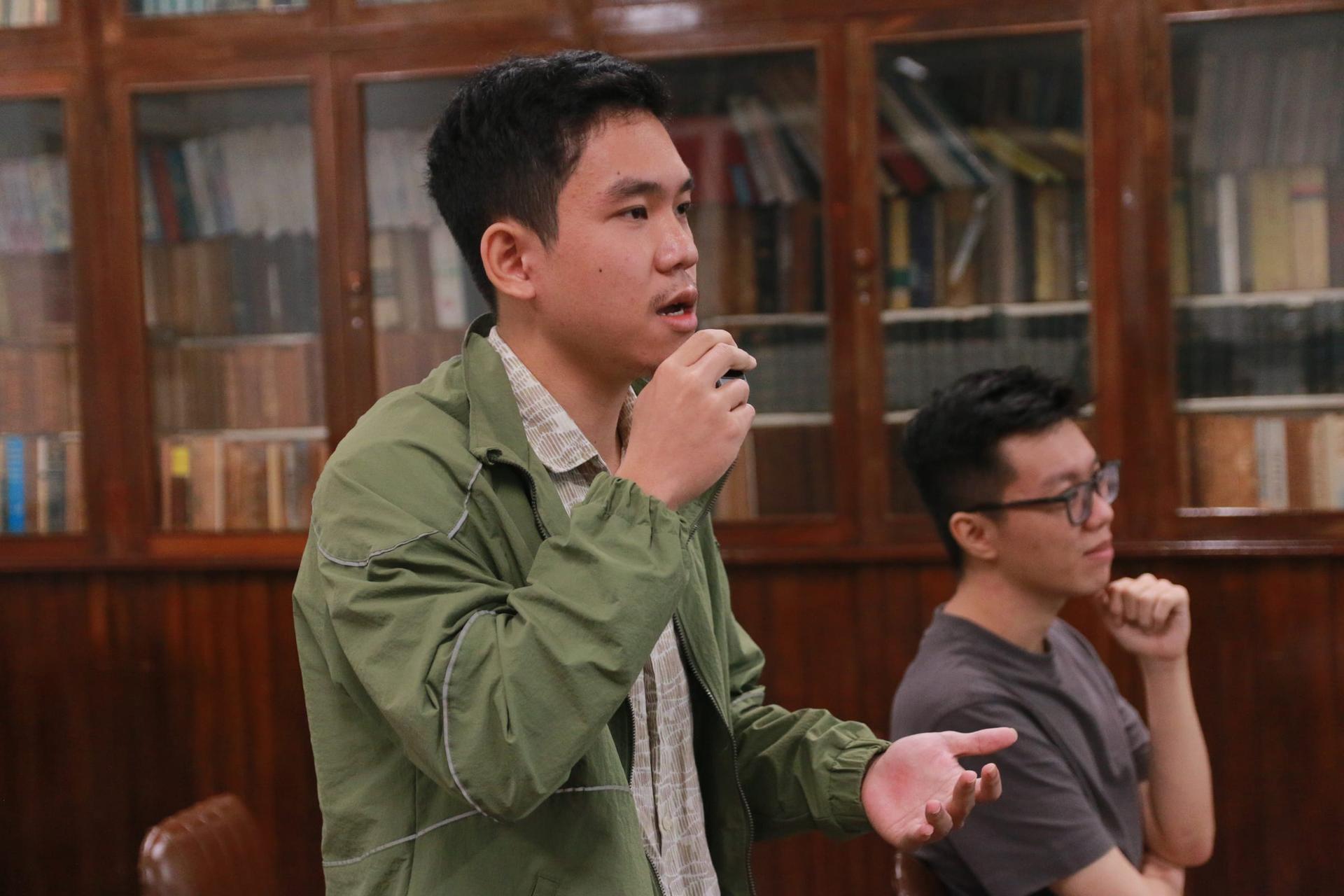 Một người nghe mặc áo khoác xanh rêu cầm micro đặt câu hỏi, người ngồi cạnh chống cằm lắng nghe, phía sau là tủ sách gỗ.