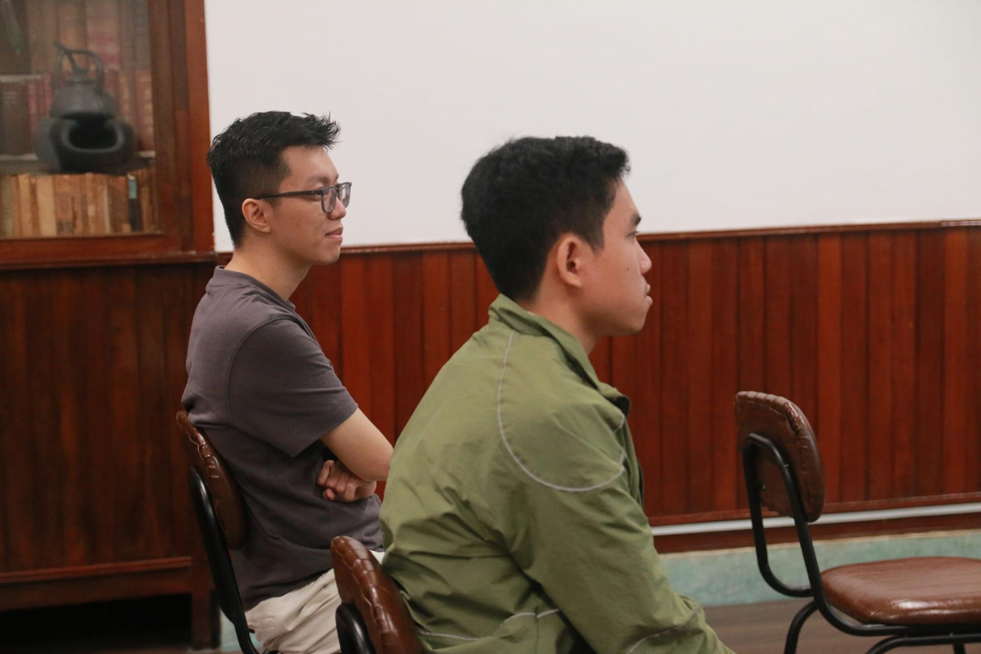 Hai người nghe chăm chú theo dõi phần trình bày, một người khoanh tay, một người mặc áo khoác xanh rêu.